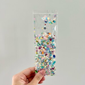 May include: A clear plastic bag filled with colorful sequins. The sequins are various shapes and sizes, including circles and flower shapes, and in shades of pink, blue, green, and silver. The bag is held in a person's hand.