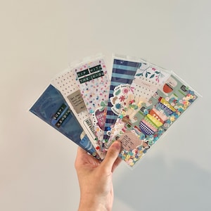 May include: A hand holding a collection of decorative bookmarks in clear plastic sleeves. The bookmarks feature various designs, including watercolor patterns, confetti, floral motifs, and text like "Busy Reading" and "Hip with the Kids."