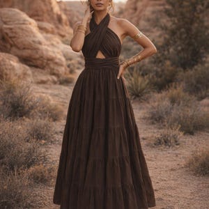 Könnte beinhalten: Braunes Maxikleid mit Neckholder-Ausschnitt, gekreuztem Oberteil und Stufenrock. Das Kleid hat einen Ausschnitt an der Taille und ist mit goldenen Armbändern und Ohrringen versehen. Aus fließendem Stoff, ideal für besondere Anlässe.