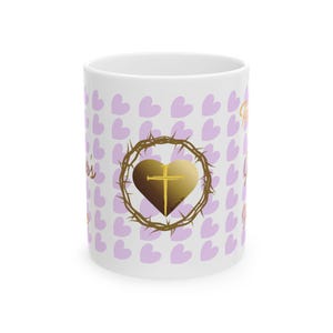 May include: White ceramic mug with a repeating pattern of light purple hearts. The design includes a gold heart with a cross, encircled by a crown of thorns. The words "Jesus" are written in gold script.