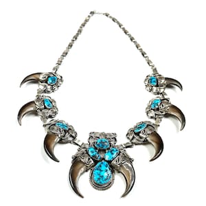 Puede incluir: Un collar de plata con colgantes de garras de oso y piedras turquesas. El collar tiene una cadena y está sobre un fondo blanco. Las garras son de color marrón oscuro.