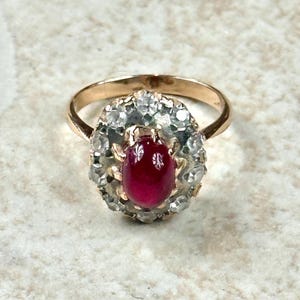 Puede incluir: Un anillo de oro con una gema roja oscura ovalada en el centro, rodeada por un halo de piedras claras más pequeñas. La banda del anillo es de oro liso. El engaste de la piedra central es ornamentado, con detalles intrincados.