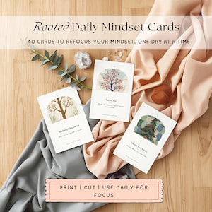 Puede incluir: Set de 40 tarjetas Rooted Daily Mindset Cards para reenfocar la mentalidad, un día a la vez. Tres tarjetas rectangulares muestran ilustraciones arqueadas de árboles y texto. Se exhiben junto a una rama de eucalipto, un cristal y tela.