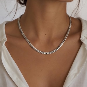 Op de afbeelding: Een zilverkleurige gourmetketting met in elkaar grijpende platte, ovale schakels. De ketting rust tegen de hals en toont een gepolijste metalen afwerking met een consistent, uniform patroon over de gehele lengte van de ketting.