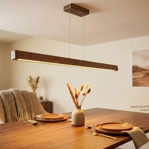 May include: A handcrafted walnut linear pendant light hangs above a wooden dining table. The rectangular light fixture is suspended by thin wires. The table is set with plates, cutlery, and a vase of dried flowers. The room has a warm, inviting atmosphere.