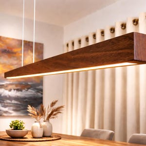 May include: A rectangular, wooden pendant light fitting with a warm, brown finish hangs above a dining table. The light has a built-in, bright, linear light source. The room features a painting and neutral-coloured curtains.