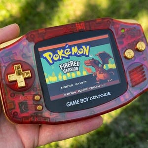 May include: A red and gold Game Boy Advance handheld console displaying the Pokémon FireRed Version game. The console has gold buttons and accents, with the game's title and a Charizard graphic visible on the screen. The console is held in a hand.