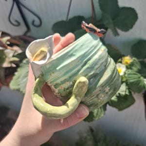Puede incluir: Una taza de cerámica hecha a mano con un diseño único. La taza presenta una combinación de colores verde claro y blanco, con un asa curvada y un detalle en forma de flor. La taza está decorada con pequeños detalles florales y una pequeña mariposa.