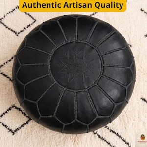 May include: A round, black leather ottoman with a geometric star design in the center. The ottoman is stitched with a contrasting thread and has a textured surface. The text "Authentic Artisan Quality" is in a yellow box.