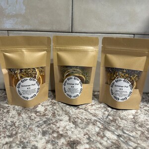 May include: Three brown paper pouches filled with dried herbs and citrus slices. Each pouch has a circular label that reads "Simmer Pot Kit" in a decorative font. The pouches are arranged on a speckled countertop against a tiled background.