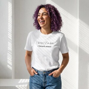 Può includere: T-shirt bianca con la scritta "Sorry I'm late, I heard music" in corsivo. La maglietta è abbinata a jeans blu. La persona sta sorridendo.