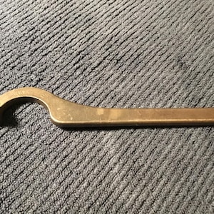 May include: A bronze adjustable hook wrench. The wrench features a curved head with a rounded end and a long, flat handle with a hole at the end. The wrench is on a textured, grey surface.