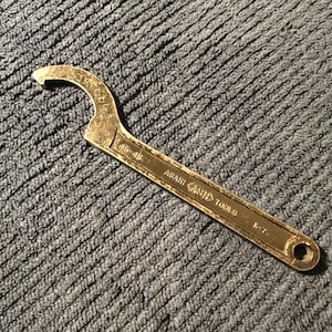 Puede incluir: Una llave inglesa de color bronce con una cabeza curva y un mango. La llave tiene grabado el texto "ASAHI (ASH) TOOLS", "15-45" y "K-7". La herramienta está sobre una superficie gris texturizada.
