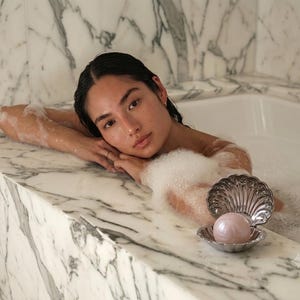 Pode incluir: Um prato de metal prateado em forma de concha contendo uma bomba de banho lisa, redonda e rosa pálido. A concha está aberta, exibindo um design interior canelado, e está posicionada sobre uma superfície de mármore branco ao lado de uma banheira.