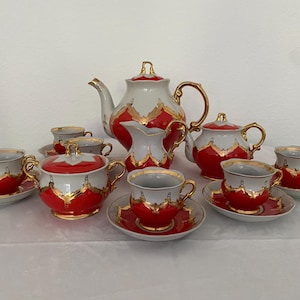 Könnte beinhalten: Ein Vintage-Teeservice mit Teekanne, Sahnekännchen, Zuckerdose und mehreren Tassen mit Untertassen. Das Set hat ein rot-weiß-goldenes Farbschema. Die Teekanne hat einen gebogenen Ausguss und einen goldenen Griff. Die Tassen und Untertassen sind kreisförmig angeordnet.