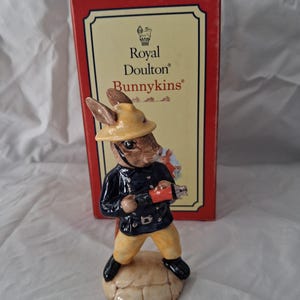 May include: A Royal Doulton Bunnikins figurine of a rabbit dressed as a firefighter, wearing a yellow helmet and holding a red hose. The rabbit is standing on a beige base, with a red and gold box in the background.