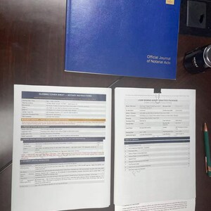 May include: A blue hardcover book titled "Official Journal of Notarial Acts" sits on a dark surface, with open documents displaying text. The documents include "Closing Cover Sheet - Notary Instructions" and "Loan Signing Agent Practice Package". A pen and a small black object are also visible.