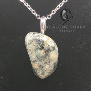 Könnte beinhalten: Halskette mit Anhänger aus poliertem, unregelmäßig geformtem Stein und silberfarbener Kette. Der Stein hat ein gesprenkeltes Aussehen mit Grautönen, Grün und Beige. Der Text "ABALONE SHARK LAPIDARY" ist sichtbar.