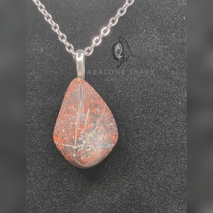 Könnte beinhalten: Eine Halskette mit Anhänger, die einen tropfenförmigen Stein mit rot-brauner Marmorierung zeigt. Der Stein hängt an einer silberfarbenen Kette. Der Text "ABALONE SHARK LAPIDARY" ist sichtbar.