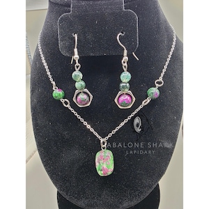 May include: A jewelry set featuring a necklace and earrings. The necklace has a pendant with a green and pink stone. The earrings and necklace have matching green and pink stone beads. The set is displayed on a black stand.