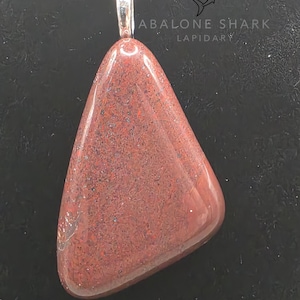 Könnte beinhalten: Ein polierter, dreieckiger Anhänger mit einem rotbraunen Stein, der an einer Silberkette hängt. Der Stein hat ein gesprenkeltes Aussehen mit dunkleren Flecken. Der Anhänger ist von Abalone Shark Lapidary.