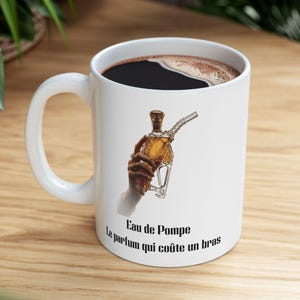 May include: White ceramic coffee mug with a black coffee fill. The mug features a graphic of a hand holding a perfume bottle with the text "Eau de Pompe Le parfum qui coûte un bras."