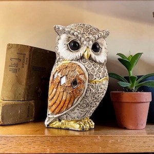 Könnte beinhalten: Eine dekorative Eulenfigur mit einem strukturierten cremeweißen Körper mit komplizierten Blumenmustern, großen schwarzen Augen, einem goldenen Schnabel und einem Flügelbereich mit orangefarbenen Akzenten und kleinen rautenförmigen Spiegeleinlagen.