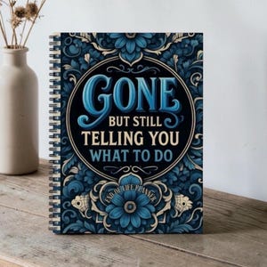 May include: A spiral-bound notebook with a dark blue floral pattern and a central circular emblem. The text reads: GONE BUT STILL TELLING YOU WHAT TO DO, with END OF LIFE PLANNER printed below. The cover features intricate blue and cream botanical motifs.