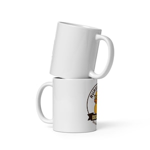 May include: Two white ceramic mugs stacked on top of each other. The bottom mug features a circular logo with a yellow rubber duck, the text "RUBBER DUCK" and "OFFICIAL". The mugs have a curved handle.
