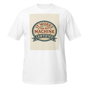 Può includere: Una t-shirt bianca a maniche corte con una grafica rettangolare centrale su sfondo beige. Il design include un emblema circolare blu scuro con il testo IT WORKS ON MY MACHINE CERTIFIED in lettere di colore rosso scuro e blu.
