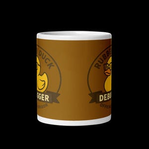 Può includere: Una tazza in ceramica bianca con un design marrone avvolgente che raffigura una papera di gomma gialla con gli occhiali. Il testo recita Rubber Duck Debugger Official Advisor in uno stile a emblema circolare. La tazza è di forma cilindrica.