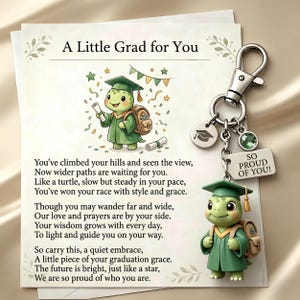 May include: A graduation-themed keychain featuring a green turtle wearing a graduation cap and gown, holding a diploma. The keychain includes a charm with the text "SO PROUD OF YOU!" and the words "A Little Grad for You".