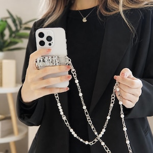 Op de afbeelding: Een witte smartphone met een rechthoekige, zilverkleurige grip met een gehamerde textuur op de achterkant. Een lange, decoratieve crossbody-riem gemaakt van afwisselende zilverkleurige metalen kettingschakels en kleine witte parels is bevestigd.