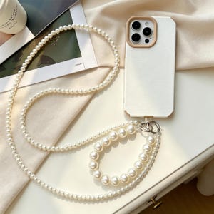May include: A white smartphone case with a tan camera frame border, featuring a long crossbody strap made of small white faux pearls and a shorter wrist strap composed of larger white faux pearls, both attached to a silver metal ring connector.