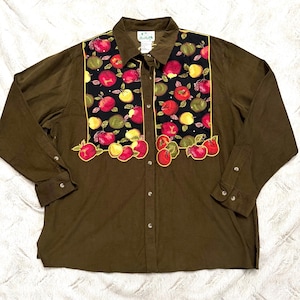 May include: A long-sleeved button-down shirt in a solid olive green color. The front features a large rectangular panel with a black background decorated with a colorful pattern of red, yellow, and green apples, outlined in bright yellow piping.
