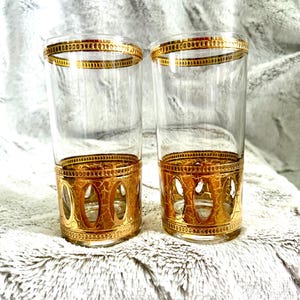 May include: Two clear glass tumblers featuring a metallic gold band at the top rim and a wider gold base section. The lower gold portion displays a textured, crackled finish with a series of vertical oval cutouts around the circumference.