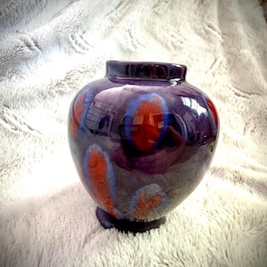 May include: A glossy, purple ceramic vase with a rounded body and a small, circular base. The vase is decorated with vibrant, abstract designs in red and blue. The vase has a smooth, reflective surface.