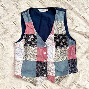 May include: A sleeveless vest featuring a patchwork design of various fabrics including red and white gingham, blue floral prints, and black star patterns. The garment has a V-neckline, four white buttons down the front, and a solid navy blue interior.