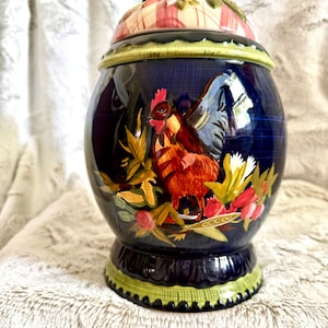 May include: A ceramic canister with a dark blue body and a lid featuring a red and white plaid pattern. The canister displays a painted rooster surrounded by corn and berries. It has a light green decorative trim at the base and the lid rim.