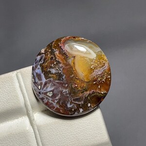 May include: A circular polished gemstone cabochon featuring a complex pattern of swirling colours including deep brown, amber, translucent white, and hints of mossy green. The surface is smooth and reflective with a high-gloss finish.