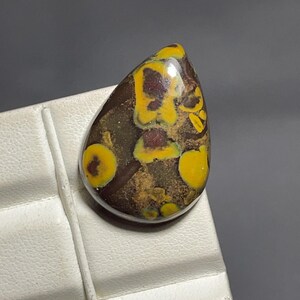 May include: A teardrop-shaped cabochon gemstone featuring a dark brown base with prominent, rounded yellow inclusions and deep red-brown spots. The stone has a smooth, polished surface with a reflective sheen, showcasing a natural, mottled pattern.