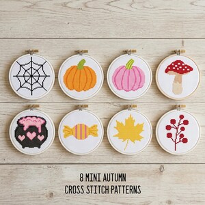 May include: Eight circular embroidery hoops containing cross-stitch patterns featuring a spider web, orange pumpkin, pink pumpkin, mushroom, cauldron with pink liquid, candy, yellow maple leaf, and red berries, with text 8 MINI AUTUMN CROSS STITCH PATTERNS.
