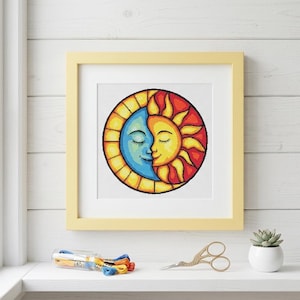 May include: A framed cross-stitch embroidery featuring a circular design of a blue crescent moon and a yellow sun with closed eyes, set against a white background. The sun and moon are surrounded by a yellow and orange border in a stained glass style.