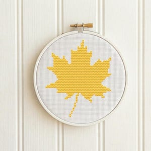 May include: A circular embroidery hoop containing white Aida fabric with a cross-stitched yellow maple leaf centred in the middle. The wooden hoop features a metal tension screw at the top, securing the fabric tightly within the round frame.