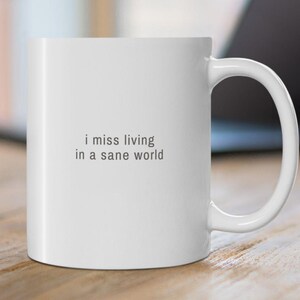 May include: White ceramic mug with the text "i miss living in a sane world" in a gray font. The mug has a curved handle and is set on a wooden surface. A laptop is visible in the background.