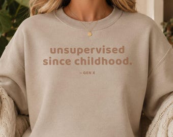 May include: A beige crewneck sweatshirt featuring the brown text "unsupervised since childhood. - GEN X" printed on the chest. The garment has long sleeves with ribbed cuffs and a ribbed neckline, paired with light blue denim jeans and white trainers.