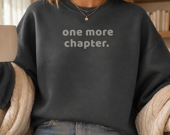 May include: A dark charcoal grey crewneck sweatshirt featuring the text one more chapter. printed in a light grey sans-serif font across the chest. The garment has long sleeves and a relaxed fit, paired with light blue denim jeans and white trainers.