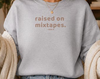 May include: A light blue crewneck sweatshirt featuring the brown text "raised on mixtapes. - GEN X" printed on the chest. The garment has long sleeves with ribbed cuffs and a ribbed neckline, worn with light blue denim jeans and white trainers.