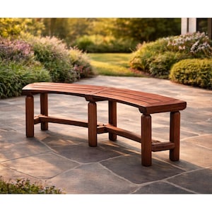 May include: A curved wooden bench with a rich brown finish. The bench has a slatted seat and is supported by sturdy, rounded legs. It is placed on a stone patio, surrounded by lush greenery, creating a natural outdoor seating area.