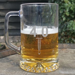 May include: A clear glass beer mug filled with beer and foam. The mug has a handle and a decorative base. The number "40" and a dagger are etched on the front of the mug. The mug is sitting on a wooden surface.